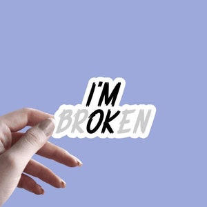 May include: A white sticker with black text that reads "I'M BROKEN" on a light purple background. The sticker is being held by a hand.