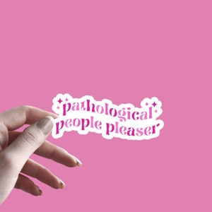 May include: A white sticker with pink text that reads "pathological people pleaser" with small stars on either side. The sticker is being held by a hand against a pink background.