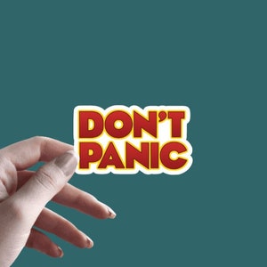 May include: A red and yellow sticker with the text "DON'T PANIC" on a white background. The sticker is being held by a hand.