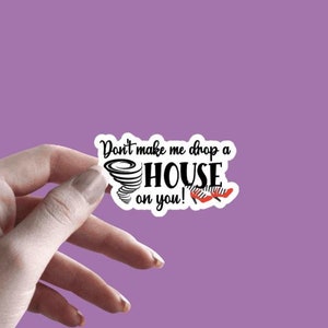 May include: A white sticker with black text that reads "Don't make me drop a HOUSE on you!" with a black and white tornado graphic and a pair of red and white shoes. The sticker is 3 inches by 1.75 inches.