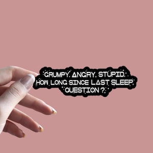 May include: A black sticker with white text that reads "GRUMPY. ANGRY. STUPID. HOW LONG SINCE LAST SLEEP? QUESTION?" is held against a pink background. The sticker is in a speech bubble shape. The sticker is from Thee Sticker Shop.