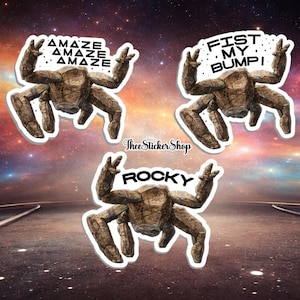 May include: Three stickers featuring a stone-like creature with raised arms. One sticker reads "AMAZE AMAZE AMAZE", another says "FIST MY BUMP!", and the last says "ROCKY". The background is a colorful galaxy.