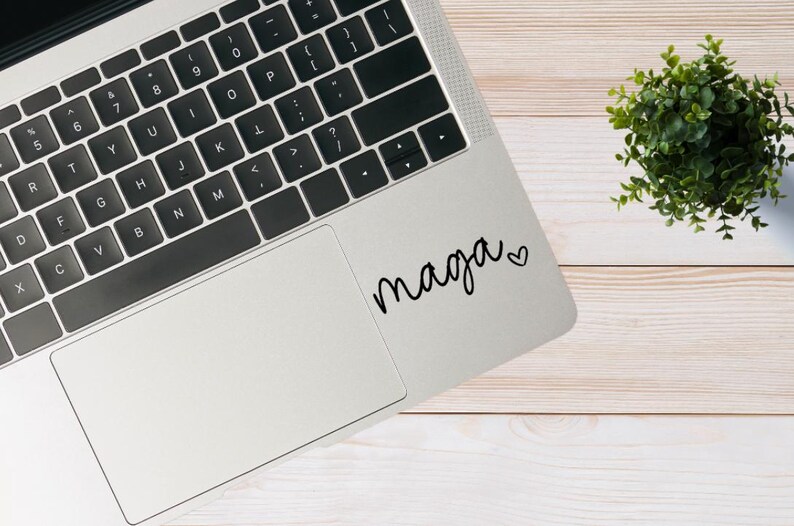 May include: A silver laptop computer with a black keyboard and a black trackpad. The laptop has a sticker on the trackpad that says "maga" with a heart symbol.