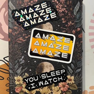 May include: A notebook with a floral and celestial design, featuring three stickers. The stickers read "AMAZE" in a repeating pattern, and "YOU SLEEP I WATCH". The notebook has a dark background with floral and moon illustrations.