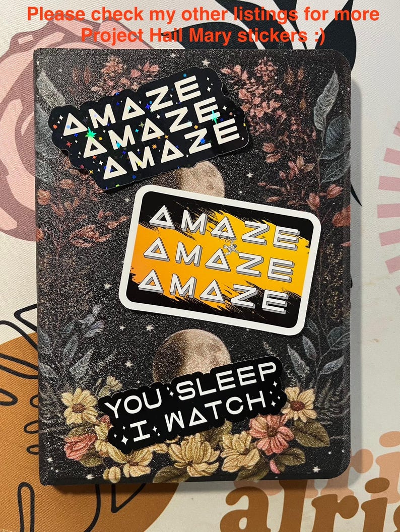 May include: A notebook with a floral and celestial design, featuring three stickers. The stickers read "AMAZE" in a repeating pattern, and "YOU SLEEP I WATCH". The notebook has a dark background with floral and moon illustrations.