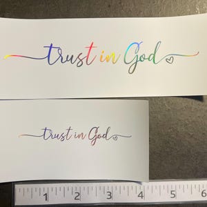May include: Two white rectangular stickers with the words "trust in God" in a cursive font. The text has a rainbow holographic effect. A small heart is next to the word "God". A ruler is at the bottom.