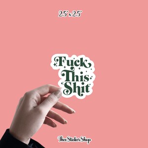 May include: A white sticker with the text "Fuck This Shit" in dark green, with decorative flourishes. The sticker is 2.5 x 2.5 inches and held by a hand against a pink background. The sticker is from Thee Sticker Shop.