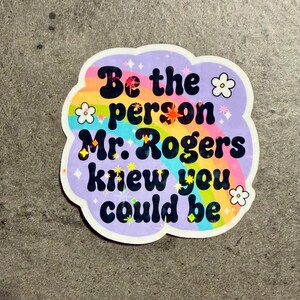 May include: A die-cut sticker with a light purple background featuring a rainbow arc and the black text "Be the person Mr. Rogers knew you could be." The design includes small white flower outlines and scattered holographic star accents throughout.