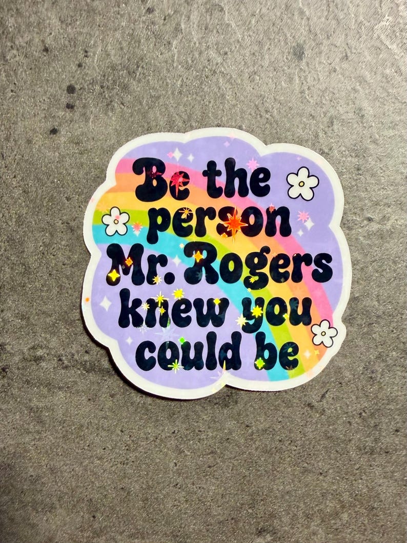 May include: A die-cut sticker with a light purple background featuring a rainbow arc and the black text "Be the person Mr. Rogers knew you could be." The design includes small white flower outlines and scattered holographic star accents throughout.