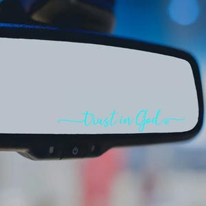 May include: A car's rearview mirror with the words "Trust in God" in a light blue script. The text is centered on the mirror's surface, with a small heart symbol at the end. The mirror is black and has control buttons at the bottom.