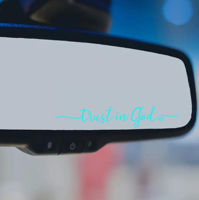 May include: A car's rearview mirror with the words "Trust in God" in a light blue script. The text is centered on the mirror's surface, with a small heart symbol at the end. The mirror is black and has control buttons at the bottom.