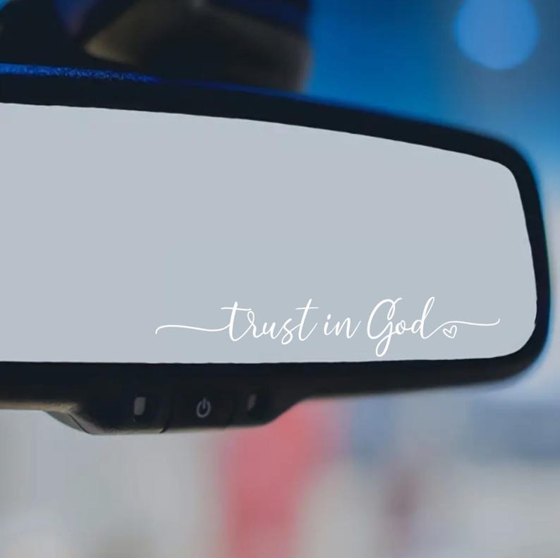 May include: A car's rearview mirror displaying the phrase "Trust in God" in elegant white cursive script. The text stands out against the mirror's grey surface. The background is blurred, suggesting a vehicle interior.