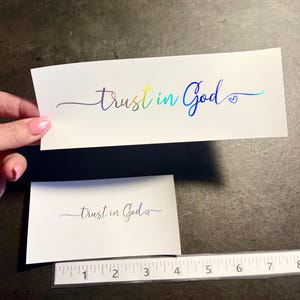 May include: Two white rectangular stickers with the text "trust in God" in a rainbow holographic font. The larger sticker is held by a hand. A ruler is placed below the smaller sticker.