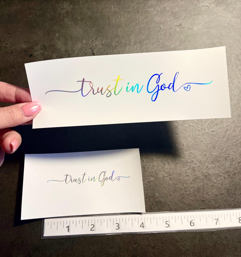 May include: Two white rectangular stickers with the text "trust in God" in a rainbow holographic font. The larger sticker is held by a hand. A ruler is placed below the smaller sticker.