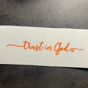 May include: A white rectangular sticker with the orange text "Trust in God" in a cursive font. A small heart is at the end of the word "God". The sticker is on a dark grey surface.