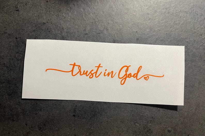 May include: A white rectangular sticker with the orange text "Trust in God" in a cursive font. A small heart is at the end of the word "God". The sticker is on a dark grey surface.