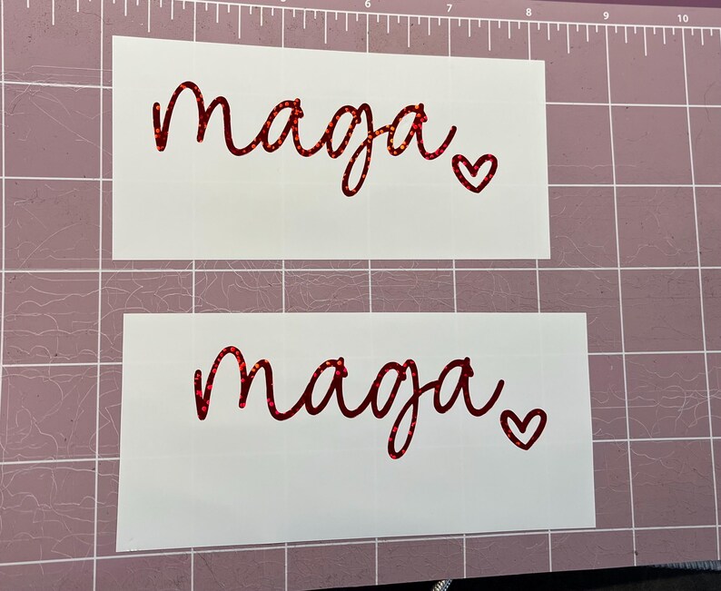 May include: Two white sheets of paper with the word "maga" written in red glitter lettering. Each sheet has a small red glitter heart next to the word.