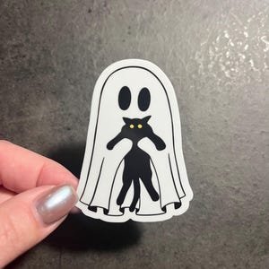 May include: A white ghost sticker with a black cat peeking out from underneath. The cat has yellow eyes.