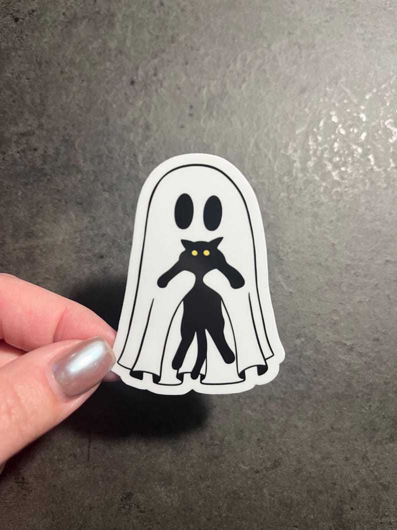 May include: A white ghost sticker with a black cat peeking out from underneath. The cat has yellow eyes.