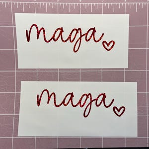May include: Two white sheets of paper with the word "maaja" written in red glitter lettering with a small red glitter heart next to it.