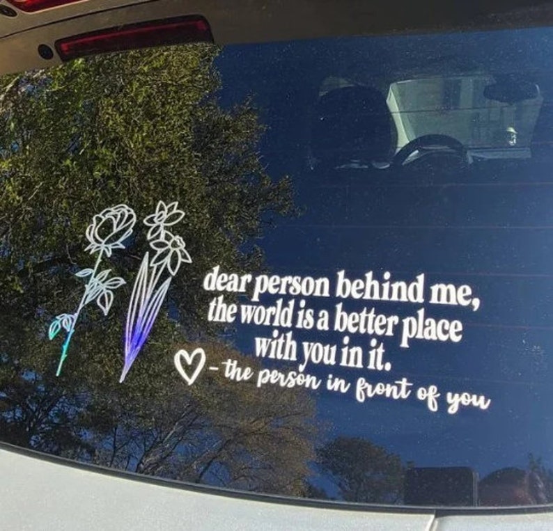 May include: A white vinyl sticker with a floral design and the text "dear person behind me, the world is a better place with you in it. - the person in front of you".