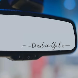 May include: A car's rearview mirror with the words "Trust in God" in elegant cursive script. The text is in black and includes a small heart at the end. The mirror is set against a blurred background.