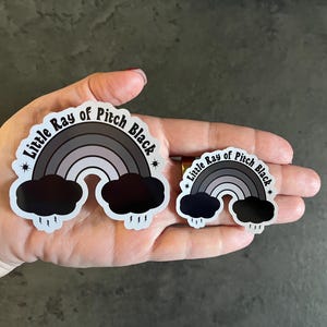 May include: Two black and white stickers featuring a rainbow and clouds. The larger sticker reads "Little Ray of Pitch Black" and the smaller sticker reads "Little Ray of Pitch Black".