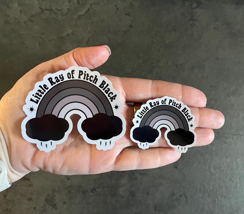 May include: Two black and white stickers featuring a rainbow and clouds. The larger sticker reads "Little Ray of Pitch Black" and the smaller sticker reads "Little Ray of Pitch Black".