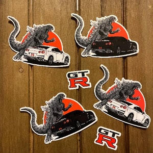 May include: Four stickers featuring a Godzilla-like monster standing on a red sun with a white or black Nissan GT-R car in front of it. The stickers also include the text "GT-R" in red and white.