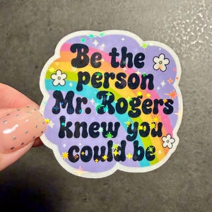 May include: A die-cut sticker with a holographic finish featuring a rainbow arc against a light purple background. The text reads, "Be the person Mr. Rogers knew you could be," in bold black letters, accented by small white flower illustrations and tiny sparkling star shapes.