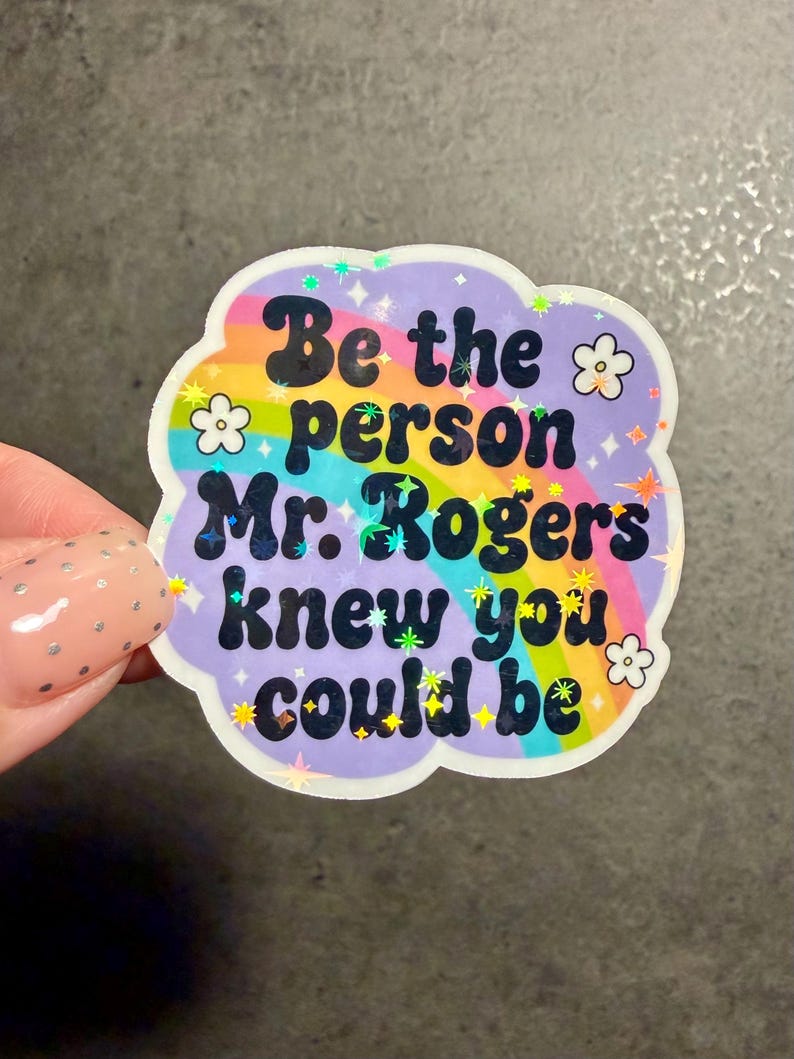 May include: A die-cut sticker with a holographic finish featuring a rainbow arc against a light purple background. The text reads, "Be the person Mr. Rogers knew you could be," in bold black letters, accented by small white flower illustrations and tiny sparkling star shapes.