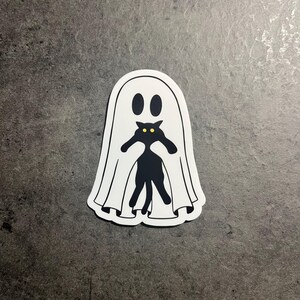 May include: A white ghost sticker with a black cat silhouette inside. The cat has yellow eyes and is peeking out from behind the ghost.