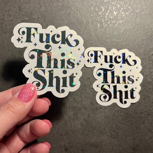 May include: Two stickers with the phrase "Fuck This Shit" in a bold, black, decorative font. The stickers have a white border and a holographic, glittery effect. One sticker is held by a hand with pink glitter nail polish.