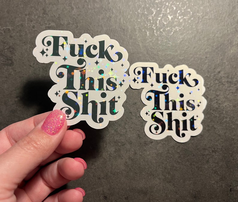 May include: Two stickers with the phrase "Fuck This Shit" in a bold, black, decorative font. The stickers have a white border and a holographic, glittery effect. One sticker is held by a hand with pink glitter nail polish.