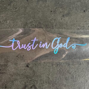 May include: A clear sticker with the words "Trust in God" in a gradient blue and purple script. A small heart graphic is next to the word "God". The sticker is held by a hand.