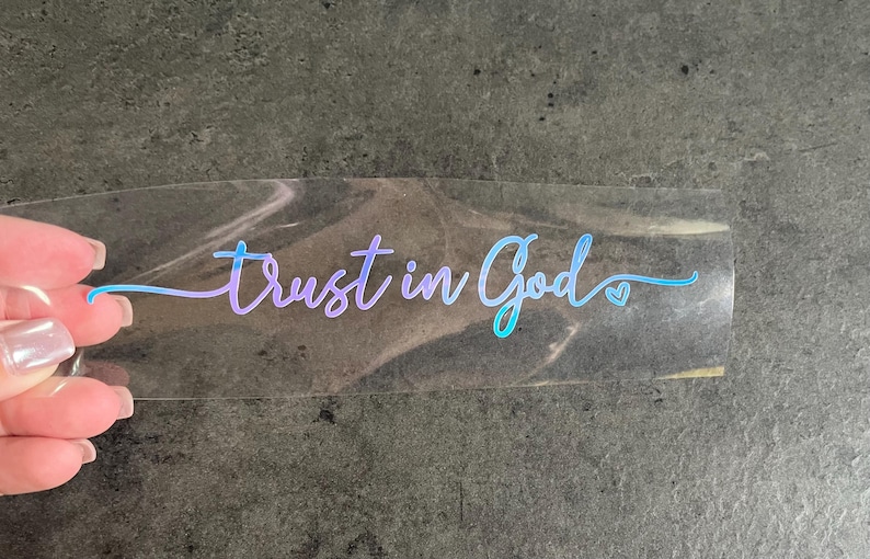May include: A clear sticker with the words "Trust in God" in a gradient blue and purple script. A small heart graphic is next to the word "God". The sticker is held by a hand.