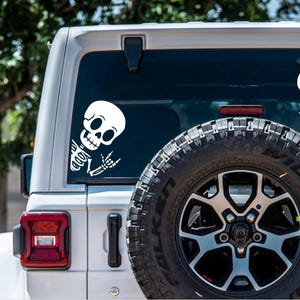 May include: A white Jeep Wrangler with a black tire and a white skeleton sticker on the back window. The skeleton is giving the rock and roll hand sign.