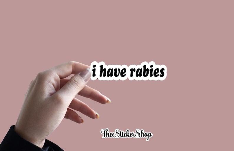 May include: A white sticker with black text that reads "i have rabies" on a pink background. The sticker is being held by a hand.