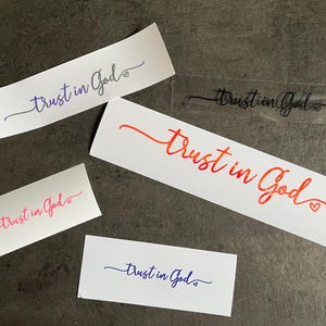 May include: A collection of stickers displaying the words "Trust in God" in different colours and fonts. The stickers are in shades of blue, pink, orange, and black. Some stickers include a heart symbol. They are arranged on a dark grey surface.