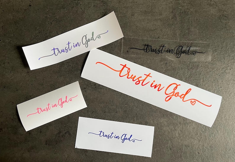 May include: A collection of stickers displaying the words "Trust in God" in different colours and fonts. The stickers are in shades of blue, pink, orange, and black. Some stickers include a heart symbol. They are arranged on a dark grey surface.