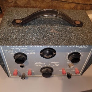 Puede incluir: Analizador de circuitos de barrido dinámico vintage, modelo 220, con una carcasa gris moteada y un asa de cuero negro. El panel frontal es gris con perillas negras, terminales rojos y texto impreso.