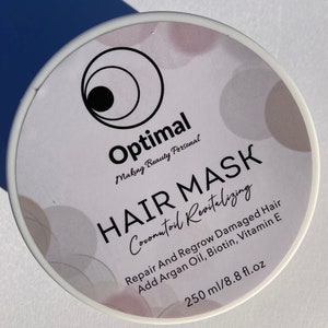 May include: White jar with a black label that reads "Optimal Making Beauty Personal HAIR MASK Coconutoil Revitalizing Repair And Regrow Damaged Hair Add Argan Oil, Biotin, Vitamin E 250 ml/8.8 fl.oz"