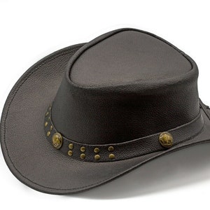 K&ouml;nnte beinhalten: Ein brauner Cowboyhut aus Leder mit breiter Krempe und einem dekorativen Band mit Metallnieten und Verzierungen.