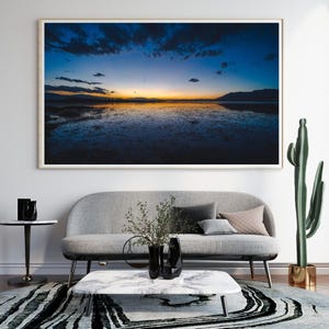 Könnte beinhalten: Ein gerahmter Landschaftsdruck zeigt einen Sonnenuntergang über dem Wasser mit blauen, orangefarbenen und gelben Farben. Der Druck hängt über einem grauen Sofa, einem Marmor-Couchtisch und einem gemusterten Teppich. Ein hoher Kaktus und schwarze Vasen vervollständigen die Szene.