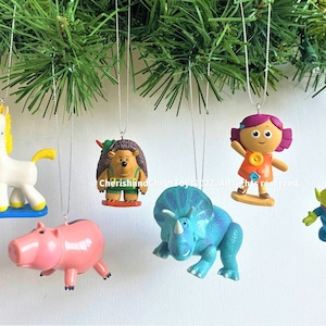 Puede incluir: Conjunto de seis adornos navideños coloridos con personajes de películas infantiles populares. Los adornos incluyen un unicornio, un erizo, una muñeca niña, un dinosaurio, un cerdo y un extraterrestre de tres ojos.