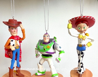 May include: Three hanging ornaments featuring Woody, Buzz Lightyear, and Jessie from Toy Story. Each character stands on a round wooden-look base and is suspended by a silver cord. The text CherishandCheerToys2025. All rights reserved. is visible.