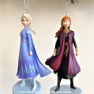 Puede incluir: Dos adornos navideños con los personajes de Frozen. Elsa lleva un vestido azul con una capa y Anna lleva un vestido morado con una capa. Ambos adornos tienen una cuerda plateada para colgar.