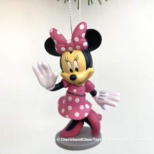Puede incluir: Un adorno navideño de Minnie Mouse. La figura lleva un vestido rosa con lunares blancos, un lazo a juego y guantes blancos. El adorno cuelga de un hilo plateado, con agujas de pino verdes en el fondo.