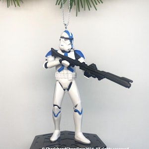 May include: A white and blue Clone Trooper figure from Star Wars, holding a black blaster rifle. The figure is a Christmas tree ornament, suspended by a silver string. The figure is mounted on a black hexagonal base.