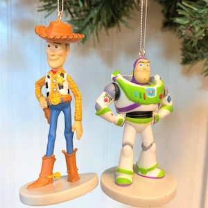 Puede incluir: Dos adornos navideños con personajes de la franquicia de películas Toy Story. El adorno de la izquierda es Woody, un muñeco vaquero con un sombrero marrón, pantalones vaqueros azules y una camisa roja y amarilla. El adorno de la derecha es Buzz Lightyear, un muñeco ranger espacial con un traje morado y verde y un casco blanco.